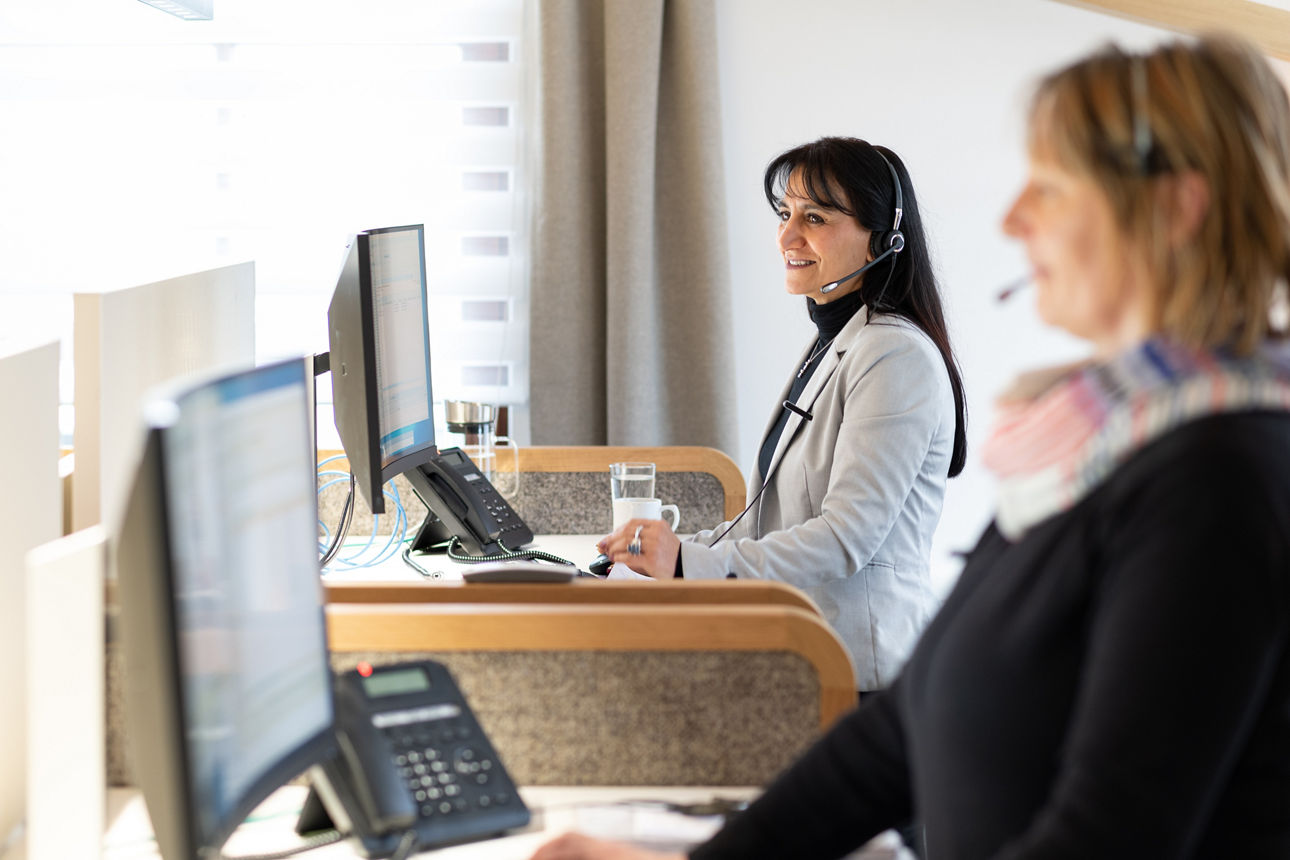 Zwei Frauen im Büro hinter Computer, blonde Frau im Vordergrund verschwommen, dunkelhaarige Frau im Hintergrund im Fokus