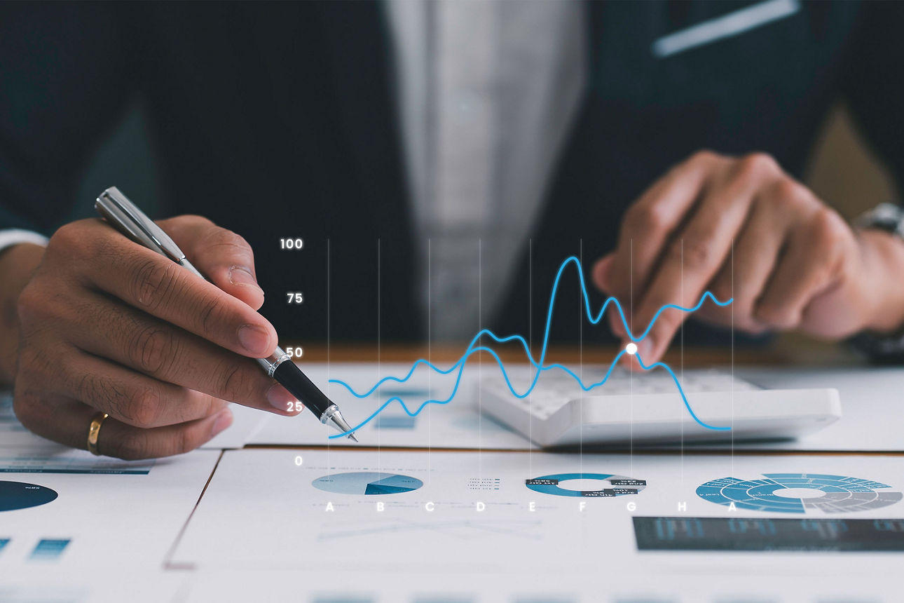 Eine Person im Anzug sitzt am Schreibtisch und hält in der einen Hand einen Stift und tippt mit der anderen Hand auf einem Taschenrechner