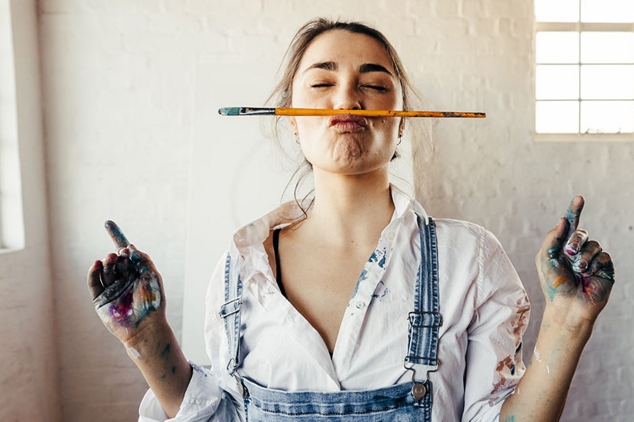Eine junge Frau steht mit Farbflecken auf der Kleidung und einem Pinsel zwischen Mund und Nase geklemmt in einem Atelier