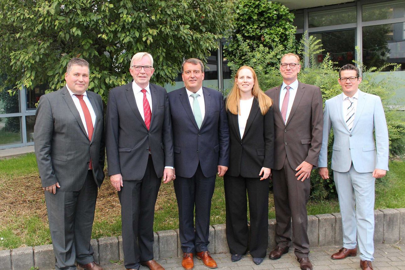 Eine sechsköpfige Gruppe, bestehend aus fünf Männern und einer Frau in Business-Kleidung, posiert lächelnd im Freien vor Büsc