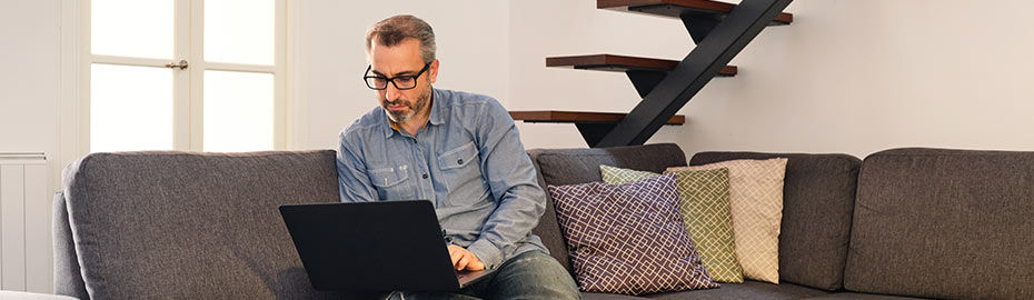 Mann sitzt bequem auf einer grauen Couch in einer modernen Wohnung und arbeitet an einem Laptop
