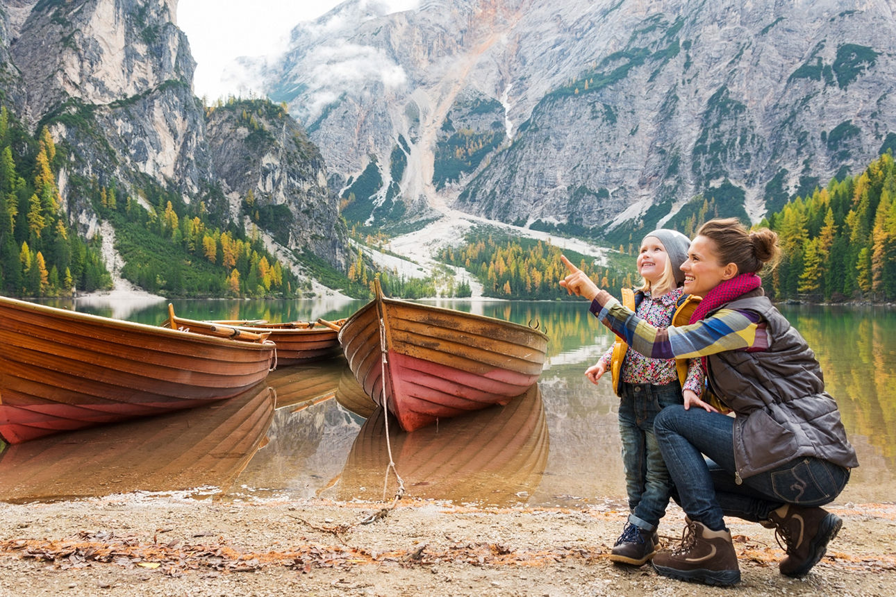 Mutter mit Kind neben Booten. Im Hintergrund ist eine Berglandschaft mit See zu sehen