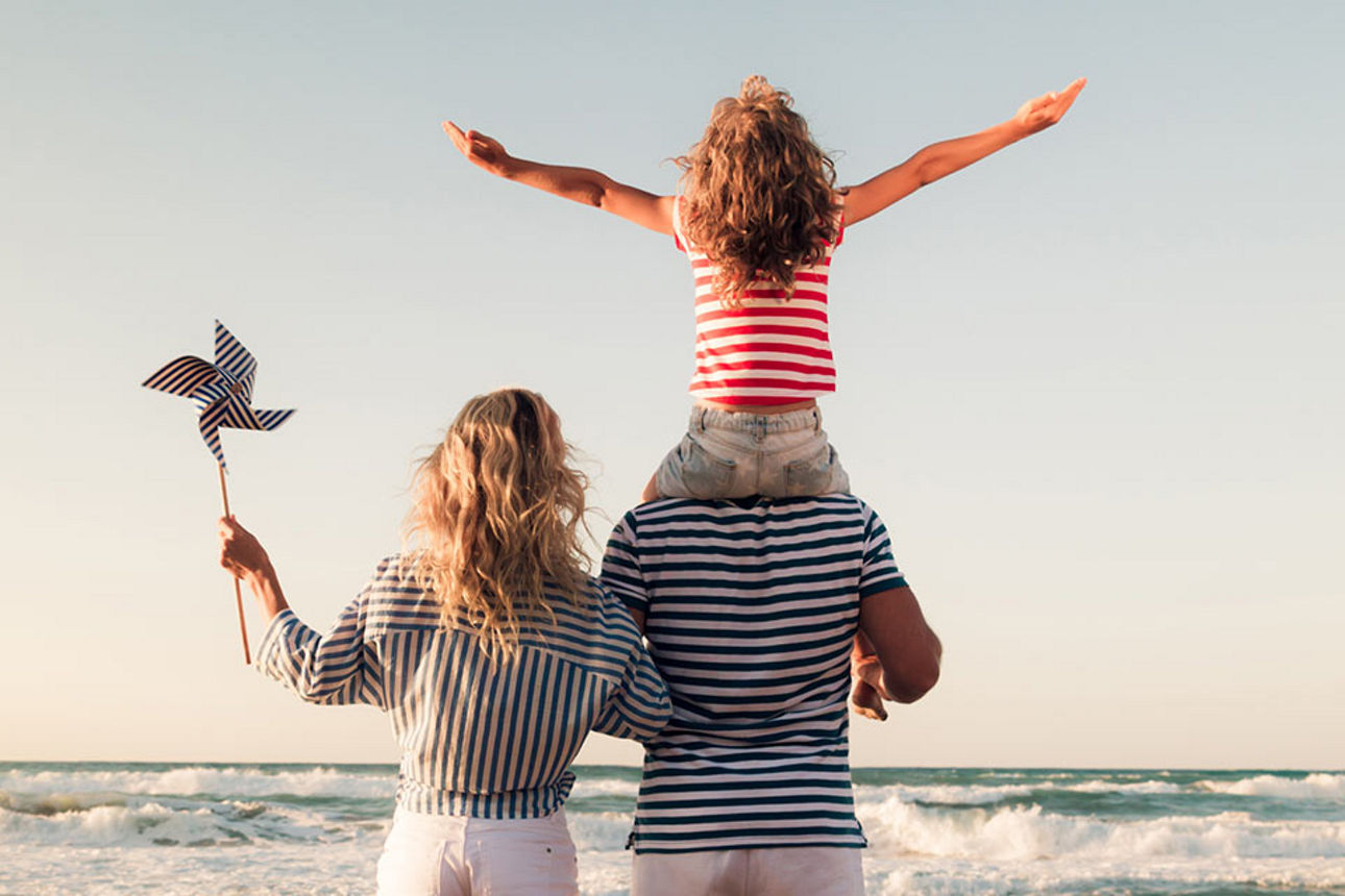 Eltern mit Tochter auf der Schulter vom Vater blicken am Strand aufs Meer