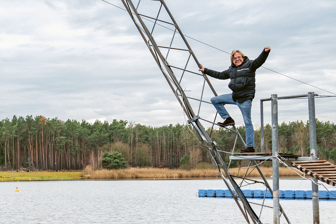 Person auf einer Wakeboard-Anlage über einem See mit Wald im Hintergrund.