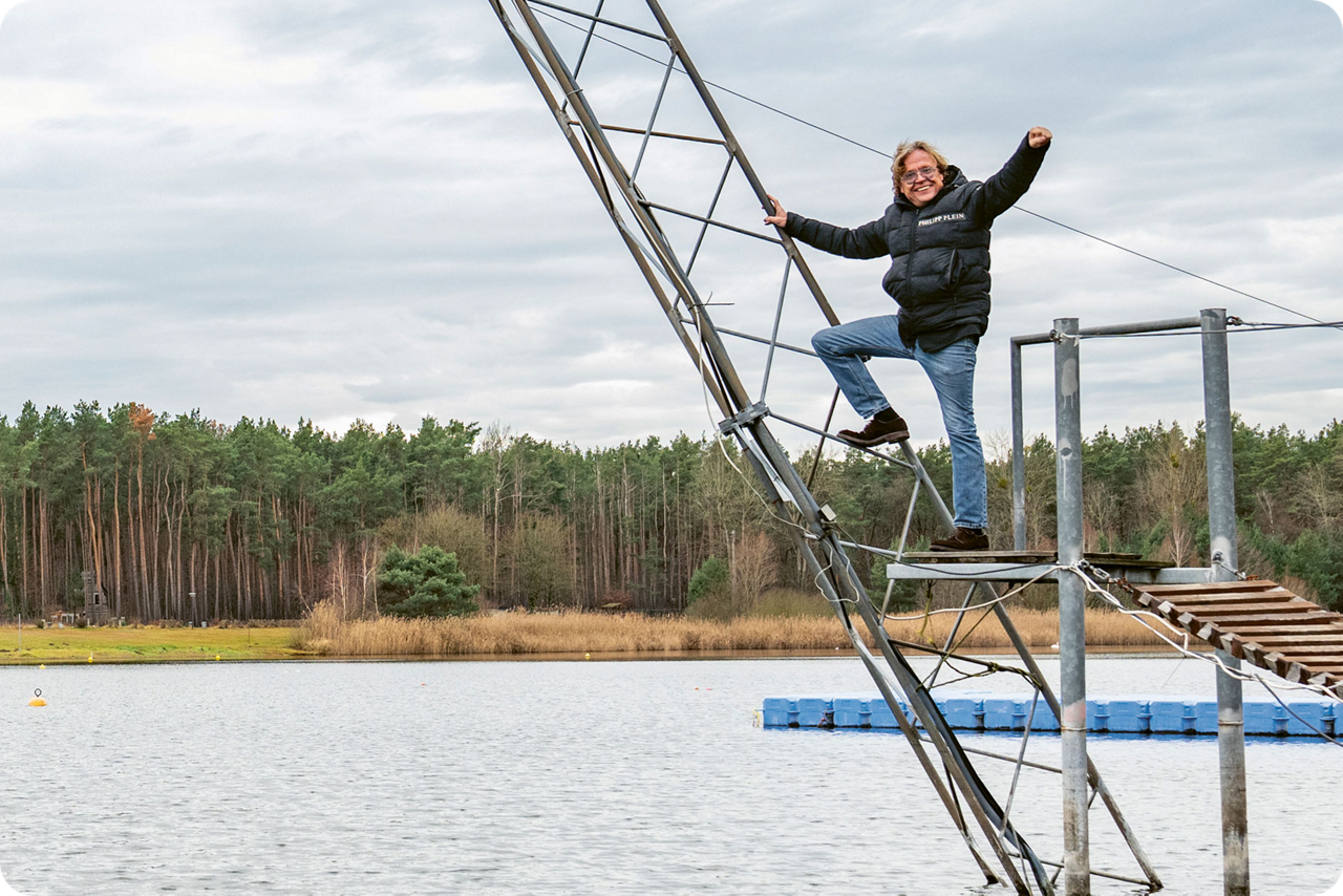 Person auf einer Wakeboard-Anlage über einem See mit Wald im Hintergrund.