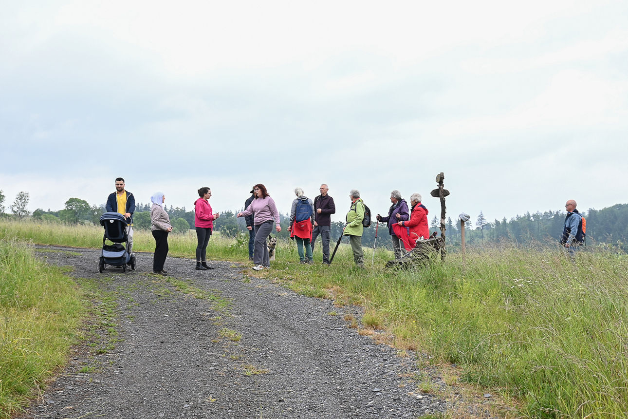 Eine Gruppe Menschen läuft um eine Kurve an einer Weggabelung. Der Weg ist geschottert. Auf der Wiese steht ein Holzwegweiser