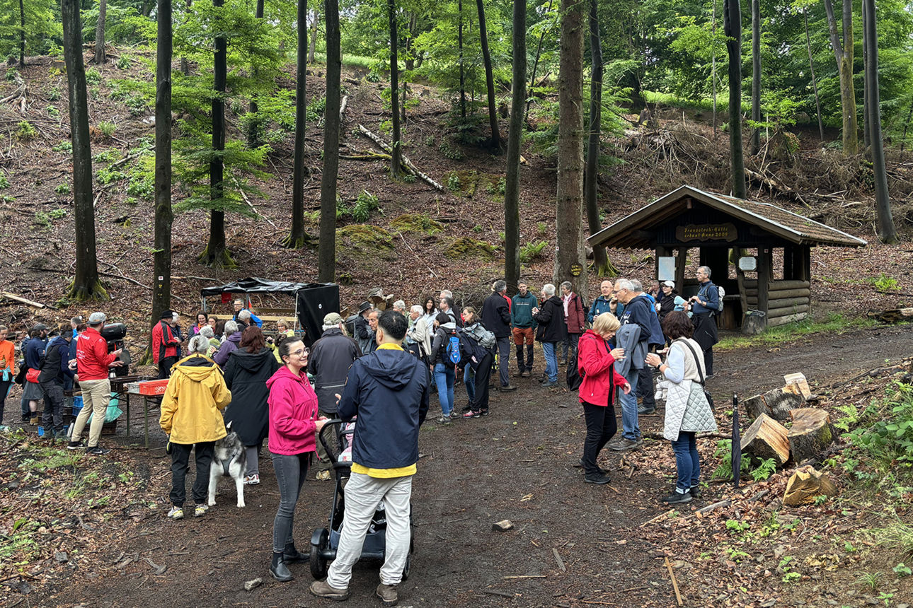 Eine Gruppe Menschen steht auf einem Platz im Wald vor einer Holzhütte. Die Personen essen, trinken und unterhalten sich.