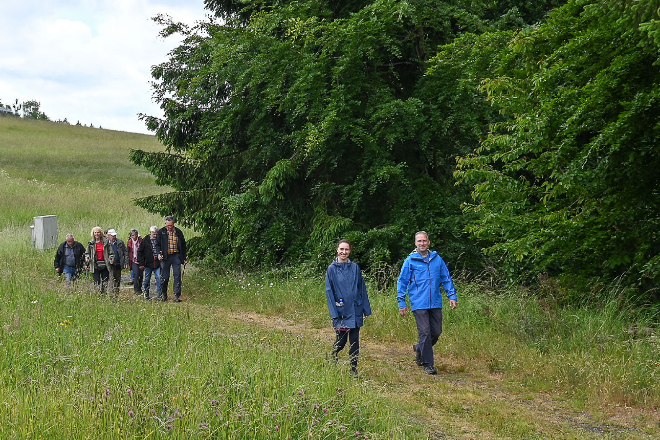 Ein Mann und eine Frau in blauen Regenjacken laufen über einen Feldweg vorbei an einem Waldstück und einer Wiese