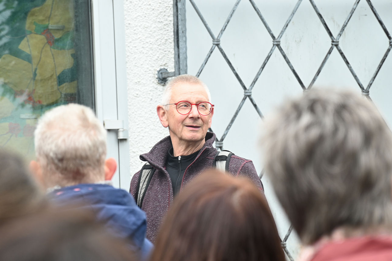 Ein Mann mit grauen Haaren und roter Brille steht vor einem vergitterten Fenster. Um ihn herum stehen viele Menschen.