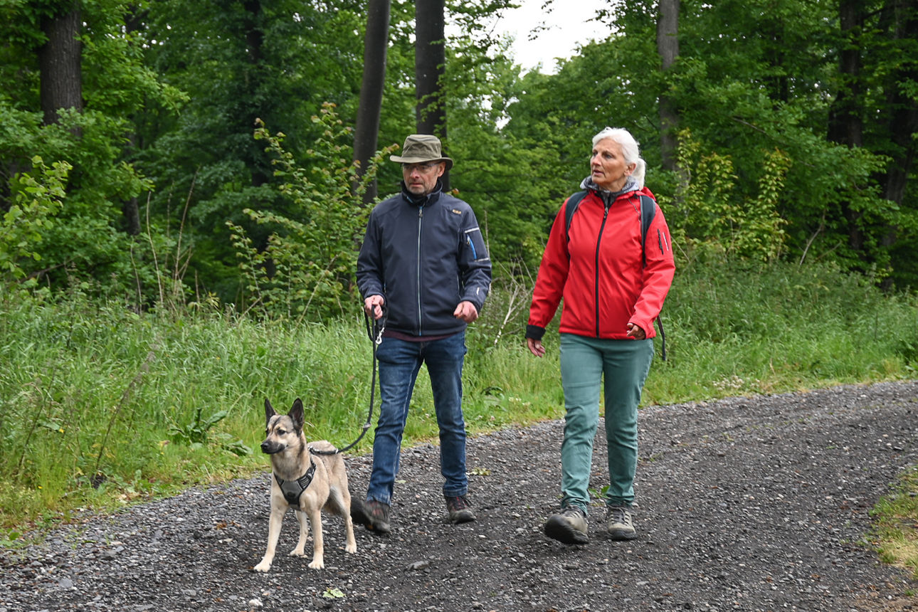 Eine Frau mit roter Jacke läuft über einen Schotterweg neben einem Mann mit Hut und Hund an der Leine
