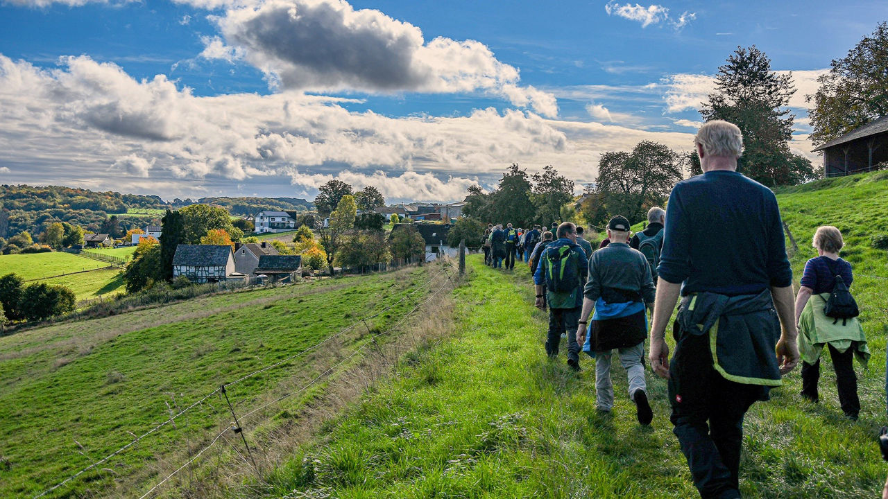 Eine Gruppe Menschen wandert bei Sonnenschein über einen Feldweg auf einer grünen Wiese in Richtung eines Dorfes