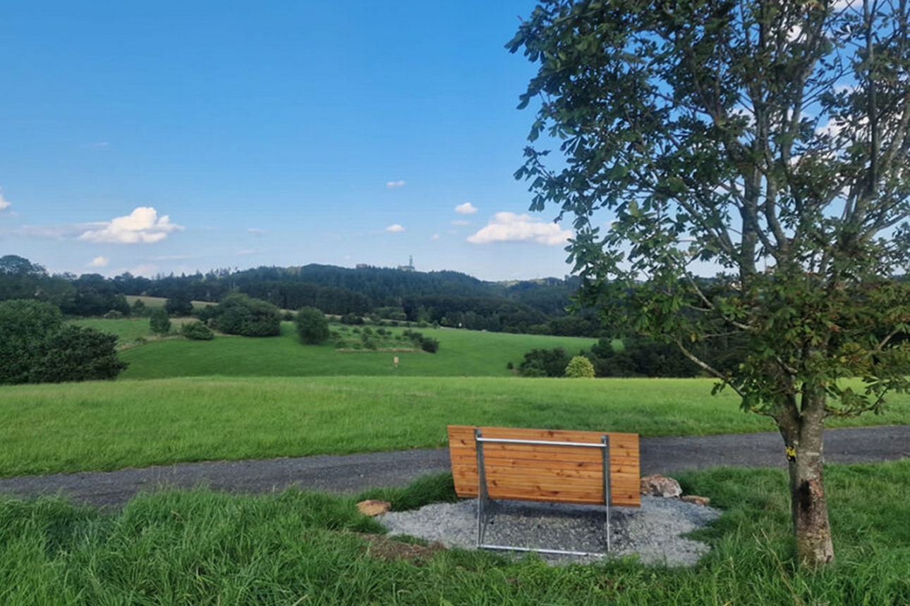 Holzbank auf einem Betonsockel mitten im Grünen neben einem Baum mit Ausblick über Wälder und Wiesen