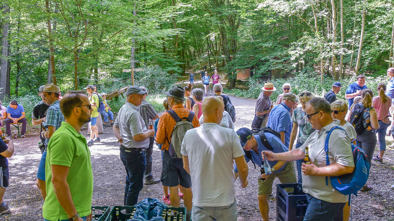 Eine große Gruppe Wandernde macht eine Pause im Schatten von Bäumen auf einer Lichtung