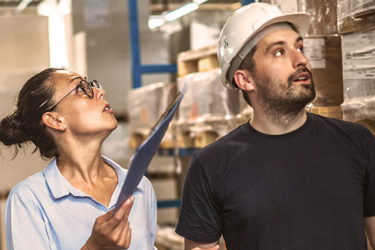 Ein Mann mit Bauhelm und eine Frau mit Klemmbrett, die etwas in einem Lagerraum suchen