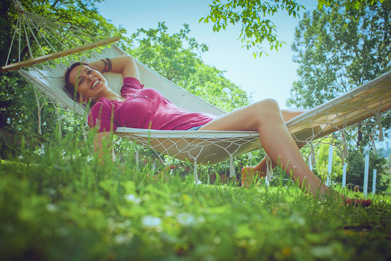 Eine junge, unbeschwerte Frau entspannt sich an einem hellen, sonnigen Tag in einer Hängematte im Garten. Sie lächelt, während sie döst und Tagträumen nachhängt. Die Aufnahme ist aus einer niedrigen Perspektive gemacht, im Hintergrund sind grüne, belaubte Bäume und ein klarer blauer Himmel zu sehen.