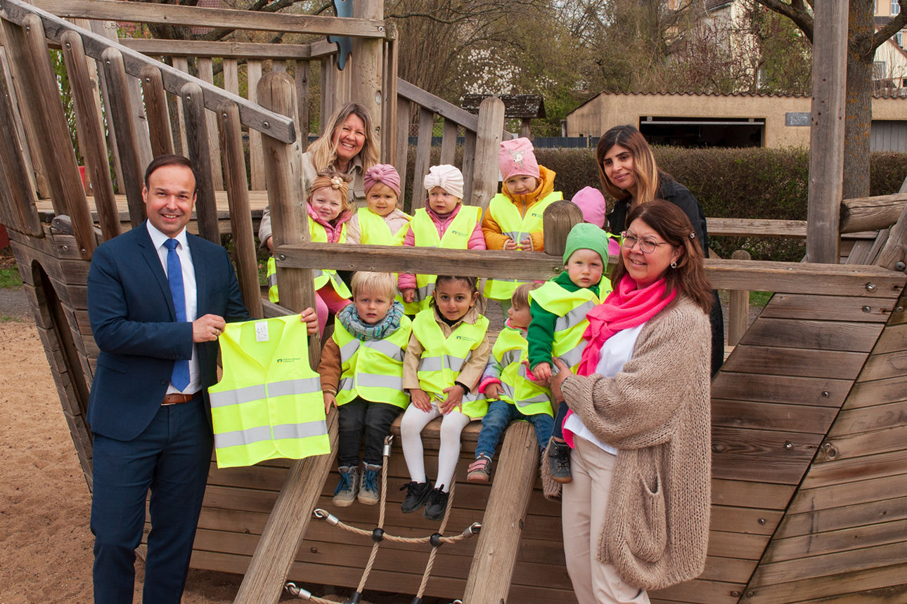 Vertriebsleiter Christian Krines übergibt die Warnwesten an die Erzieherinnen und Kinder der Mini-Kita auf Spielplatz am Main