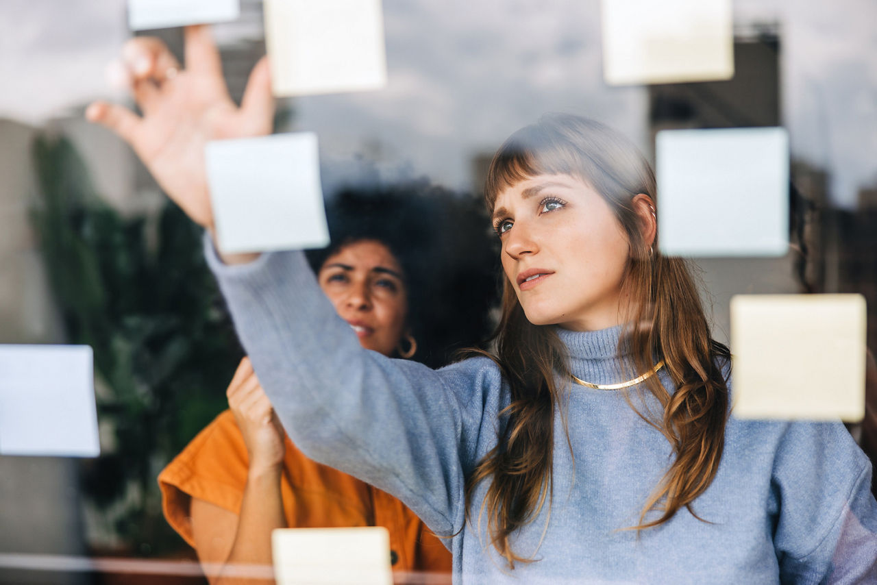 Zwei Frauen arbeiten konzentriert an Klebezetteln vor einer Glasscheibe.