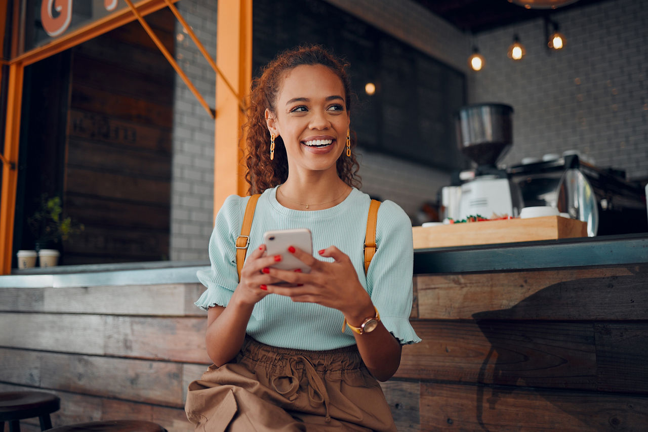 Eine junge Frau sitzt lächelnd mit ihrem Handy in der Hand und schaut auf ihre BankingApp.