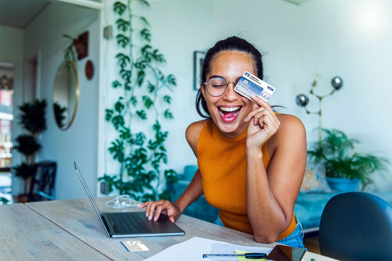 Eine Frau sitzt lachend am Schreibtisch und hält die girocard Visa Debit in der Hand.