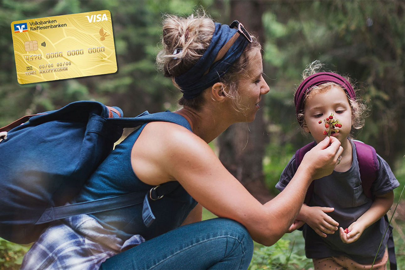 Kleines Mädchen riecht an einer Wildblume welche ihre Mutter ihr hinhält. Beide sind in Outdoorkleidung. Goldcard sichtbar