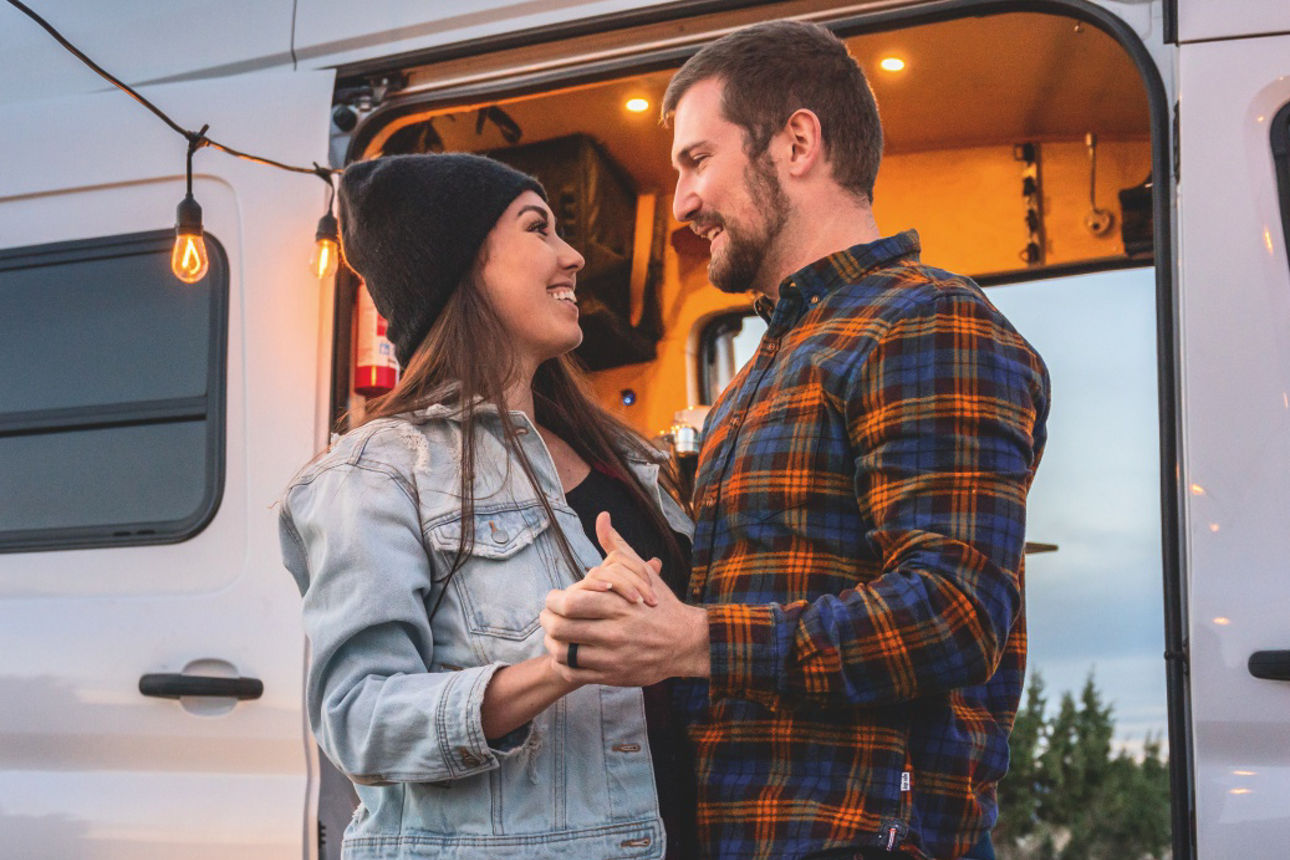 Zwei Personen stehen vor einem geöffneten Campervan mit Lichterkette und halten eine Tasse in den Händen