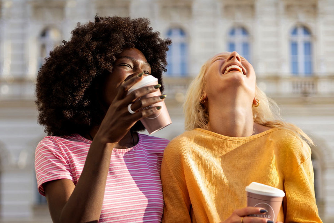 Zeigt zwei Frau mit Kaffee in der Hand, die herzhaft lachen