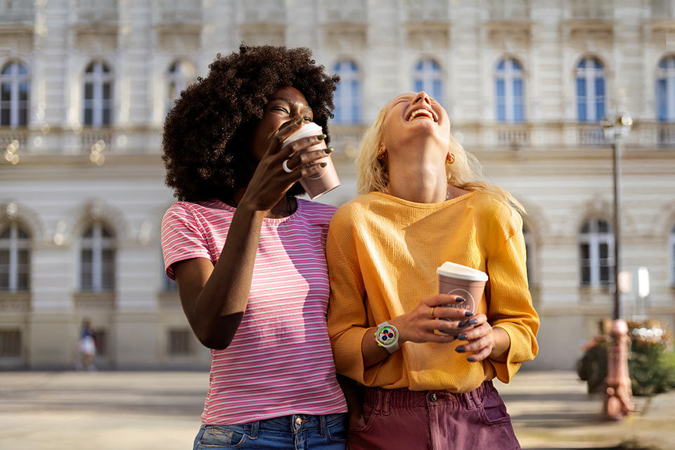 Zwei lachende junge Frauen mit Coffe-to-go-Bechern in der Hand, beide tragen Wearables wie Bezahlring und Uhr