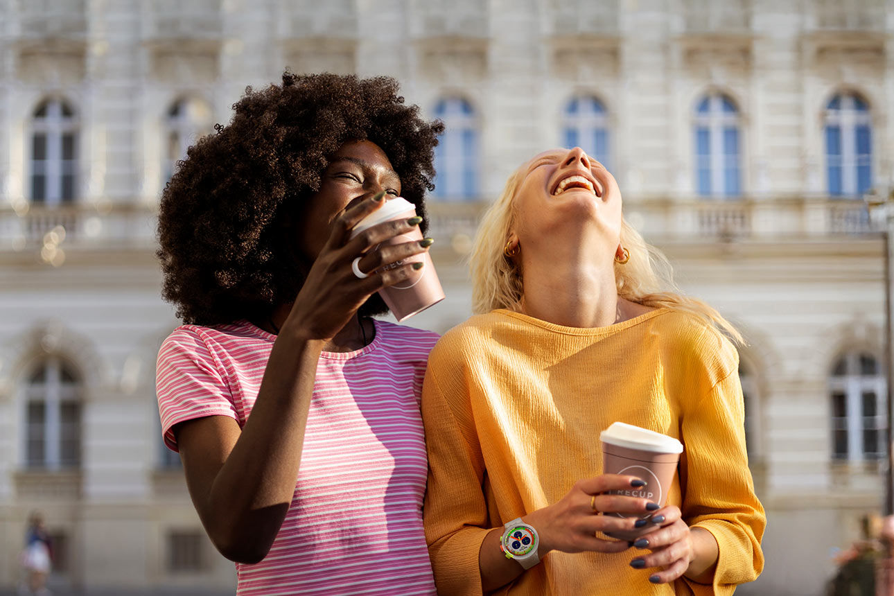 Zwei lachende junge Frauen mit Coffe-to-go-Bechern in der Hand, beide tragen Wearables wie Bezahlring und Uhr