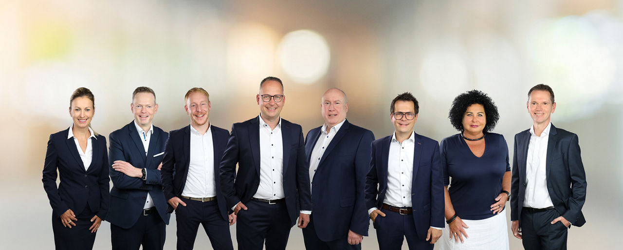 Eine Gruppe von elf Personen in Businesskleidung steht nebeneinander vor einem unscharfen Hintergrund.