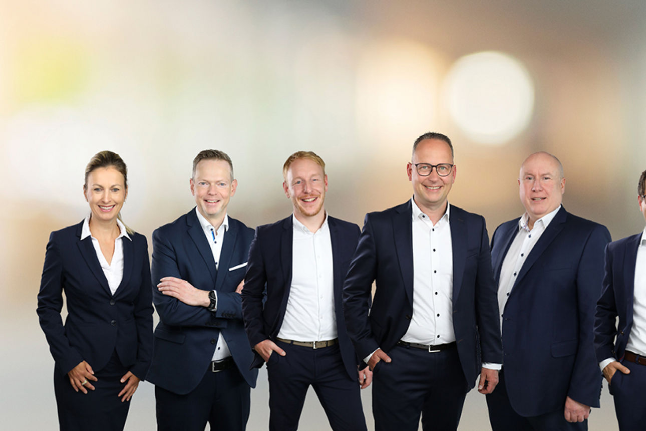 Eine Gruppe von neun Personen in Businesskleidung steht nebeneinander vor einem unscharfen Hintergrund.