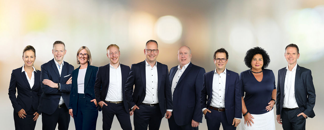 Eine Gruppe von elf Personen in Businesskleidung steht nebeneinander vor einem unscharfen Hintergrund.