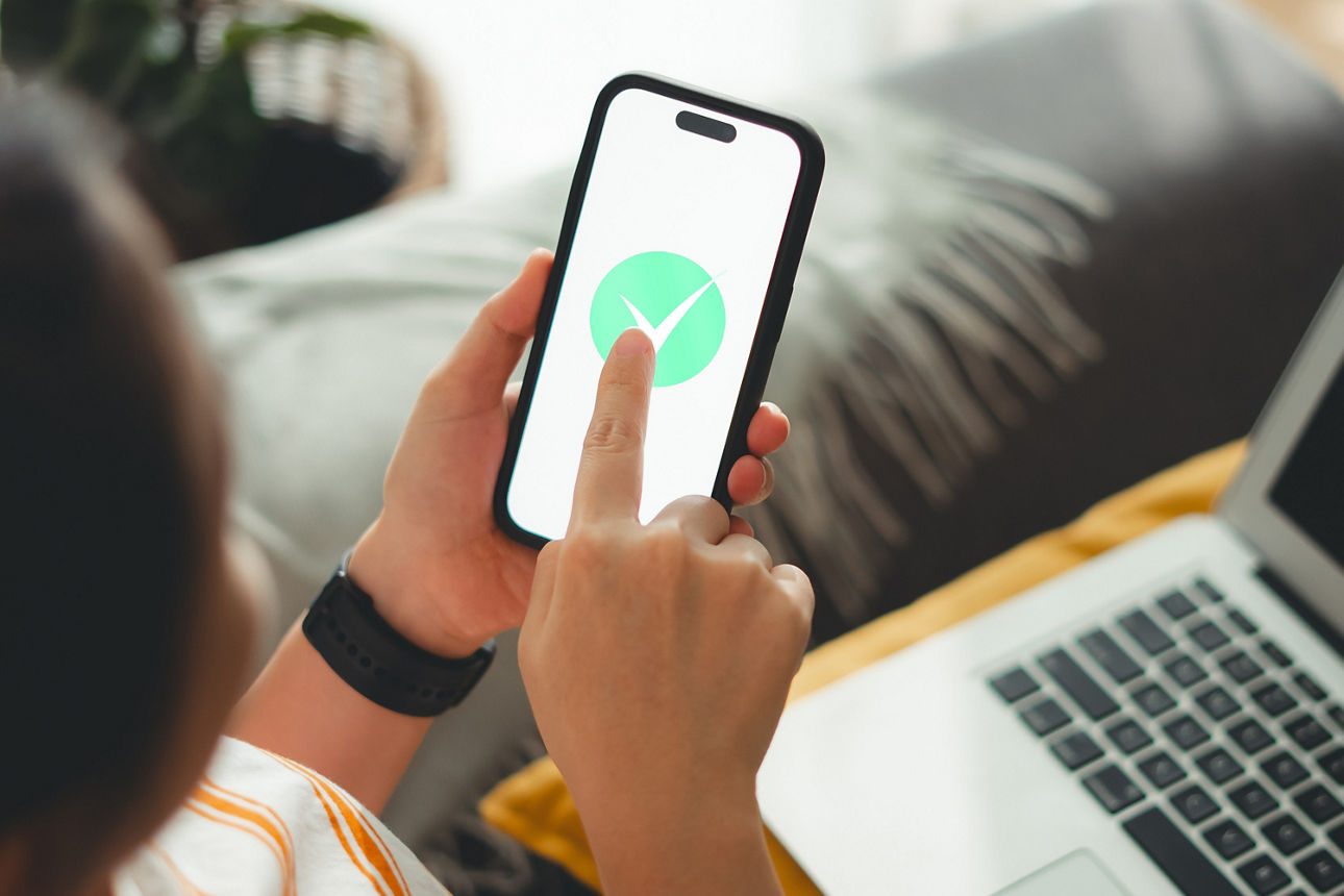 Person tippt mit Finger auf grünen Haken auf Smartphone-Bildschirm, im Hintergrund ist ein Laptop