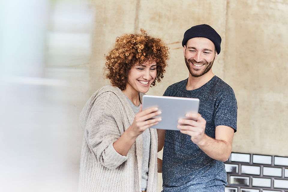 Junge Frau und junger Mann halten gemeinsam ein Tablet und schauen freudig darauf