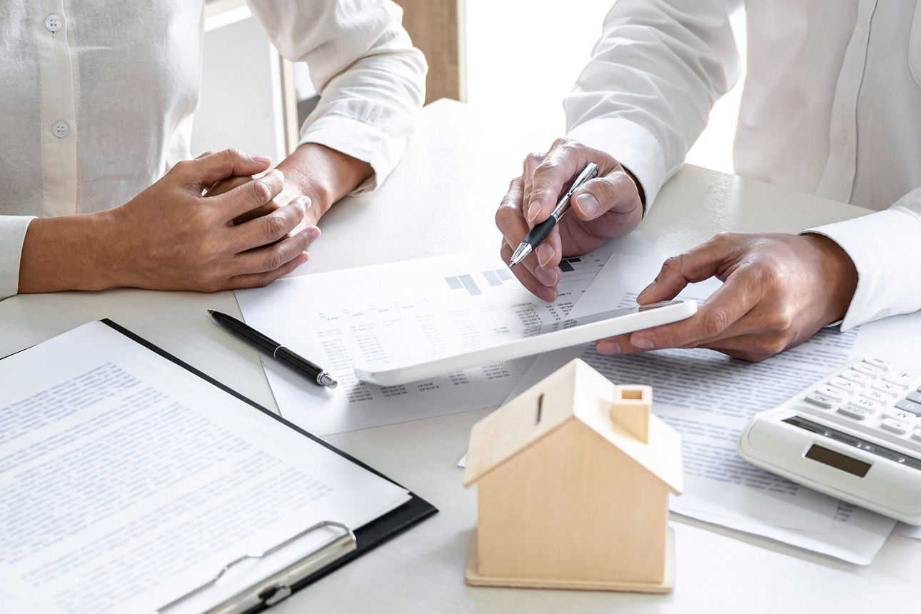 Zwei Personen in weißem Hemd die an einem Tisch sitzen und Dokumente die auf dem Tisch liegen. Ein kleines Holzhaus auf dem Tisch