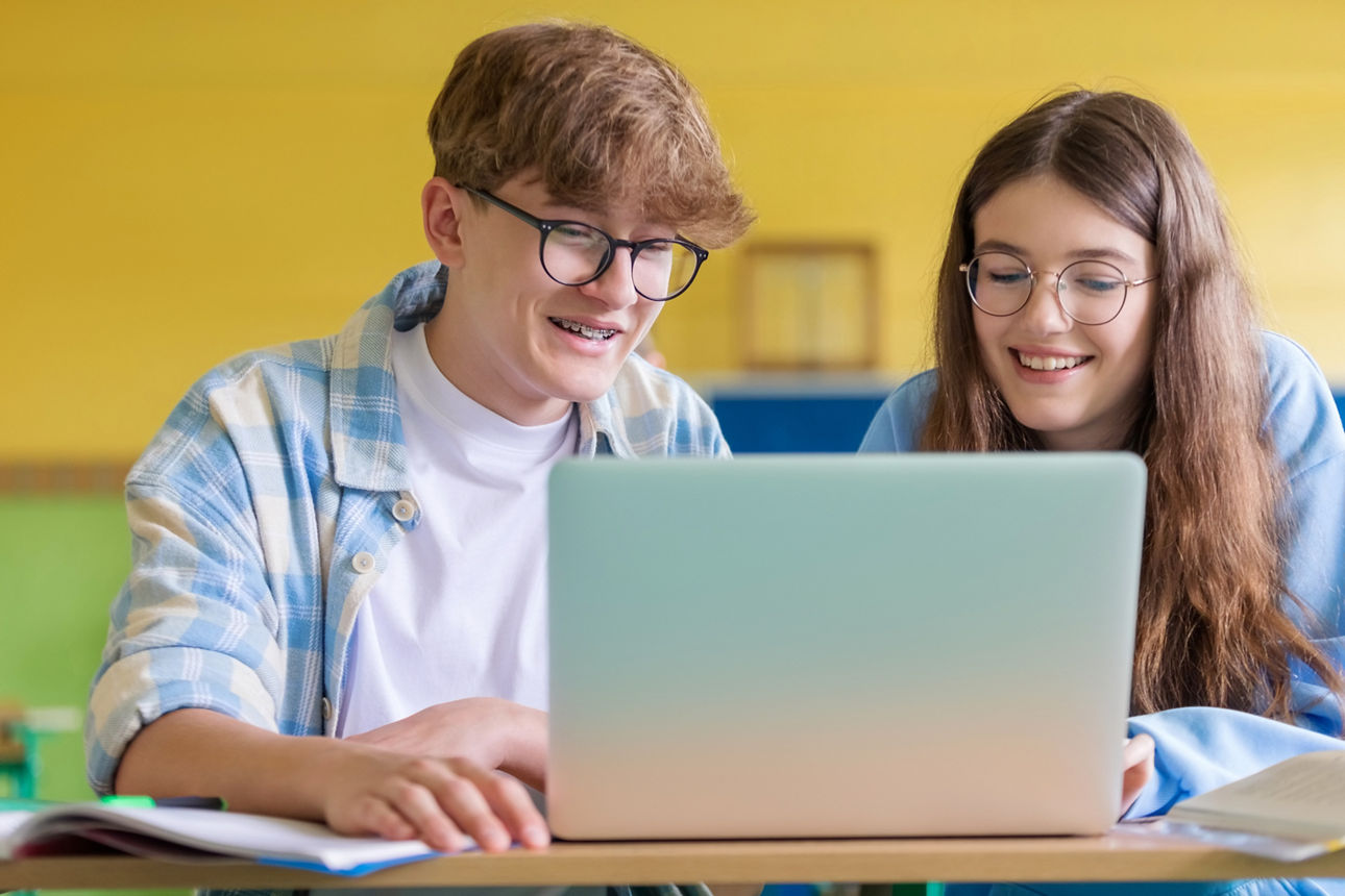 Schüler sitzen im Klassenzimmer am Pult und reden während des Unterrichts. Ein Mädchen und ein Junge arbeiten gemeinsam an einem Laptop.