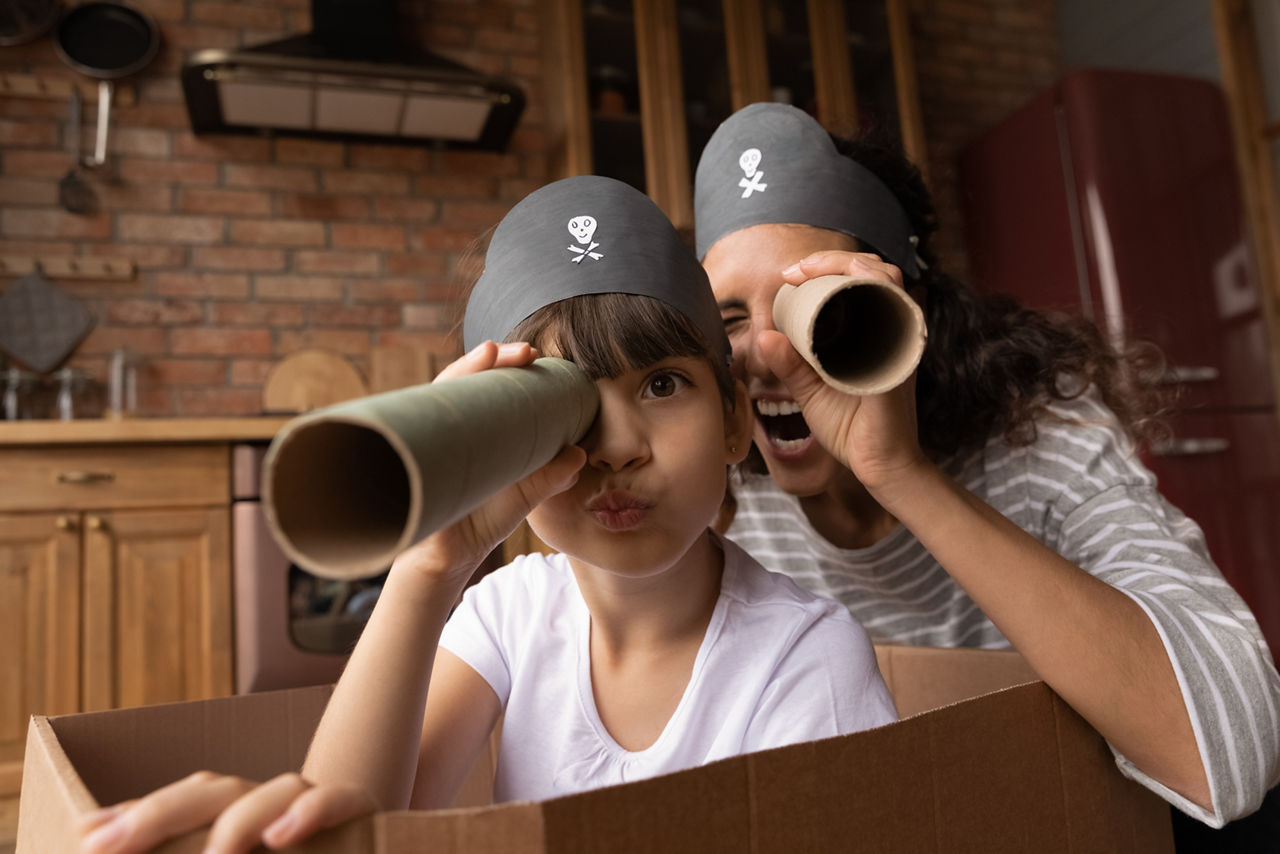 Eine Mutter und ihr Kind mit Piratenhüten spielen in einem Pappkarton und blicken durch Fernrohre aus Papp