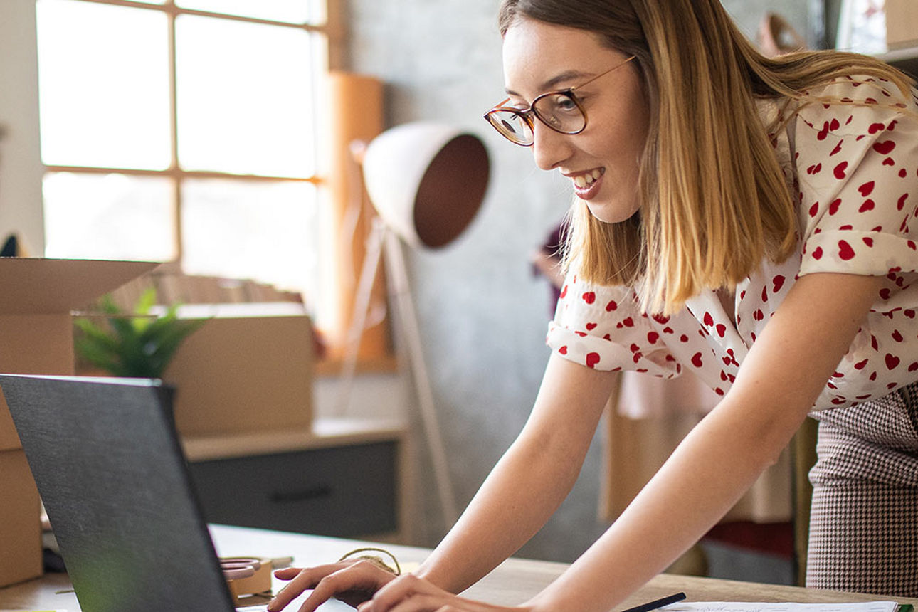 Eine Frau steht an ihrem Arbeitsplatz und tippt etwas in ihren Laptop.