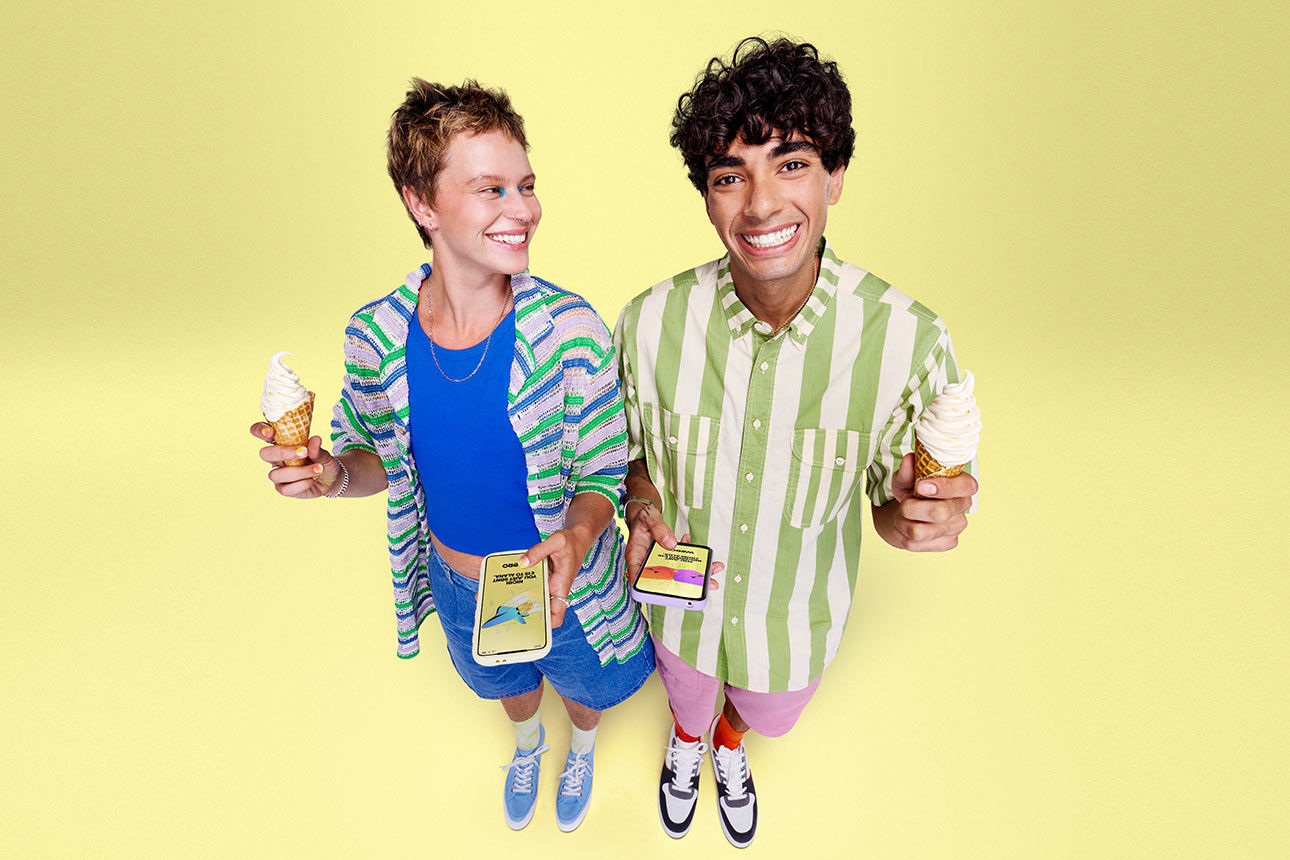 Zwei Personen stehen mit ihren Smartphones und einem Eis in der Hand lächelnd vor einem gelben Hintergrund.