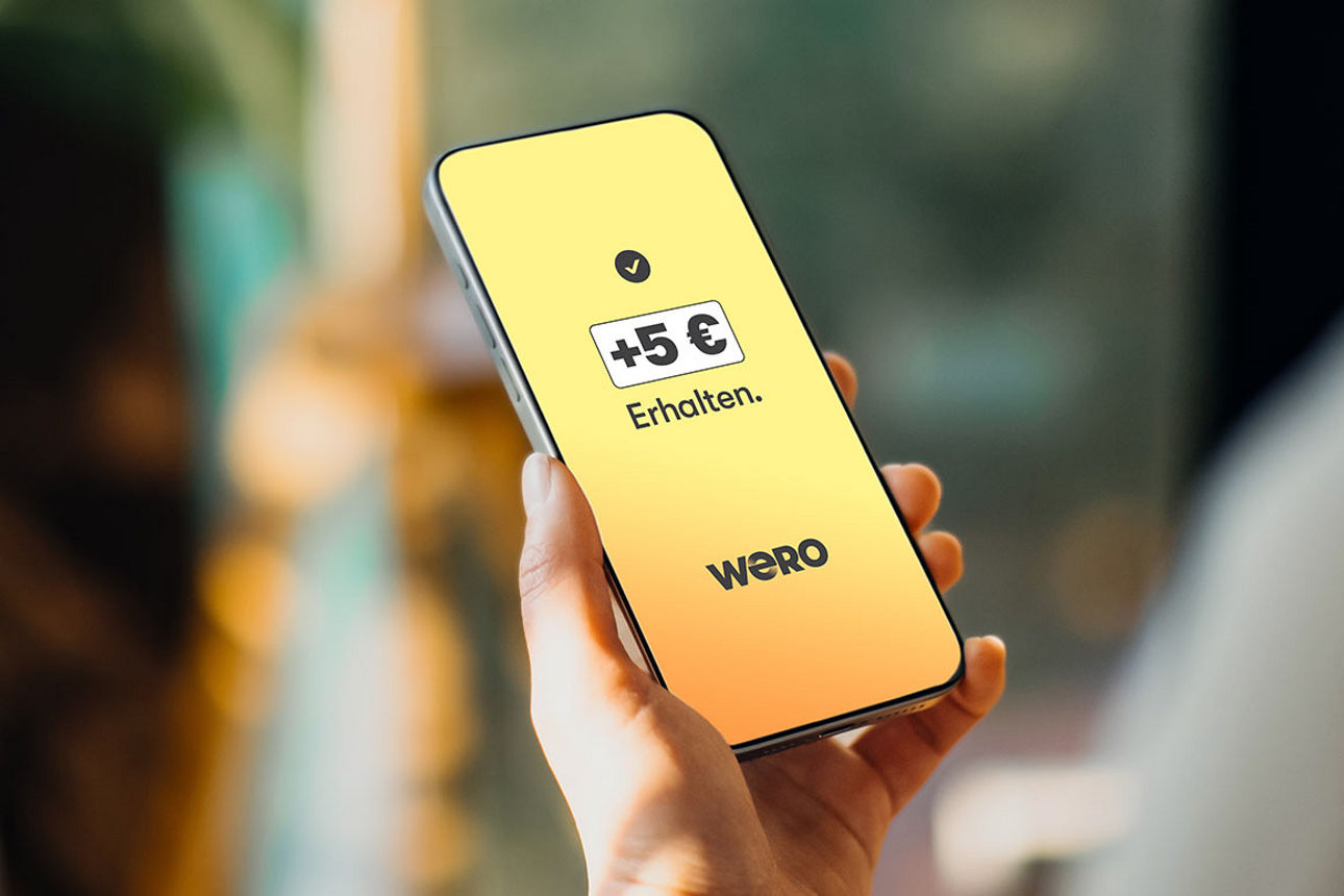 Eine Hand hält ein Handy mit einer Grafik von Wero und der Information, dass Neukunden 5 € erhalten. 