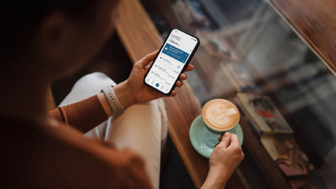 Fräu hält in linker Hand ihr Smartphone mit Banking App und in rechter Hand eine Tasse Cappucino