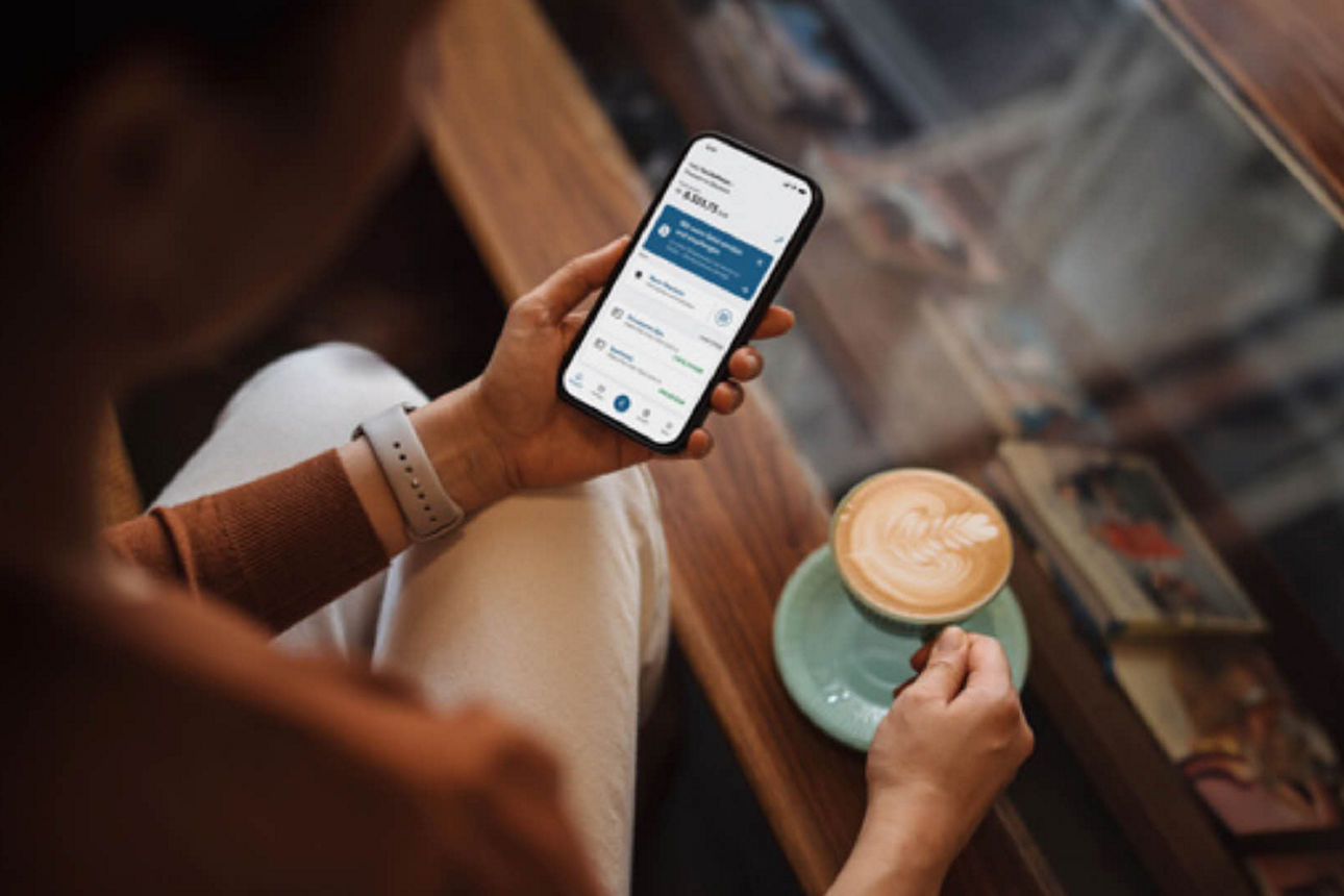 Fräu hält in linker Hand ihr Smartphone mit Banking App und in rechter Hand eine Tasse Cappucino