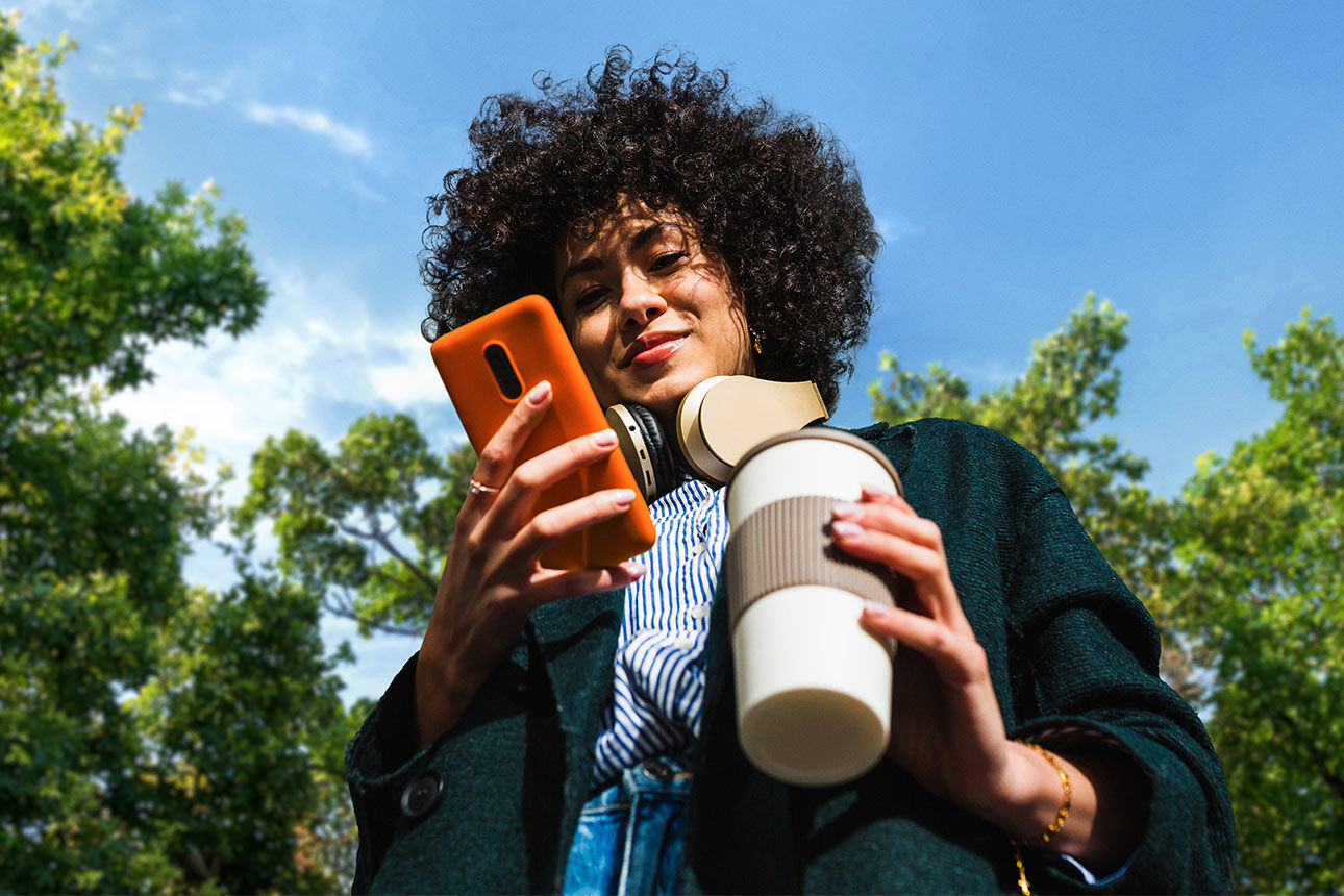 Eine Frau schaut lächelnd auf ihr Handy und hält dabei einen Kaffeebecher in der Hand.