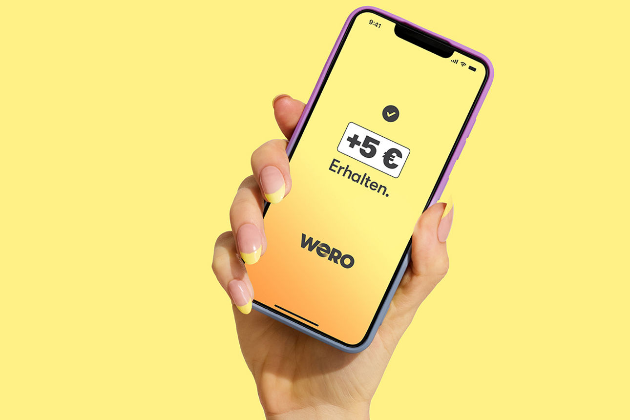 Weibliche Hand hält Smartphone mit wero-App und den Hinweis: + 5 Euro