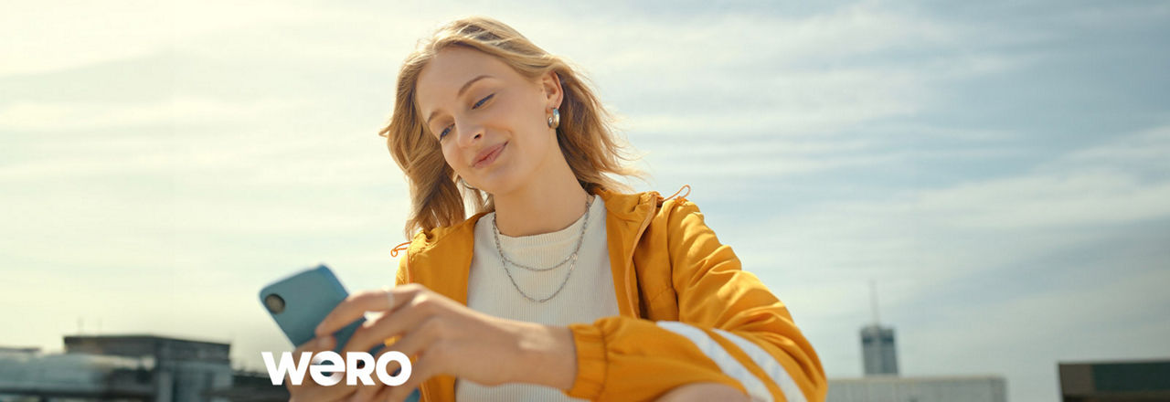 Frau mit Handy in der Hand und blauem Himmel im Hintergrund. Logo von Wero abgebildet.