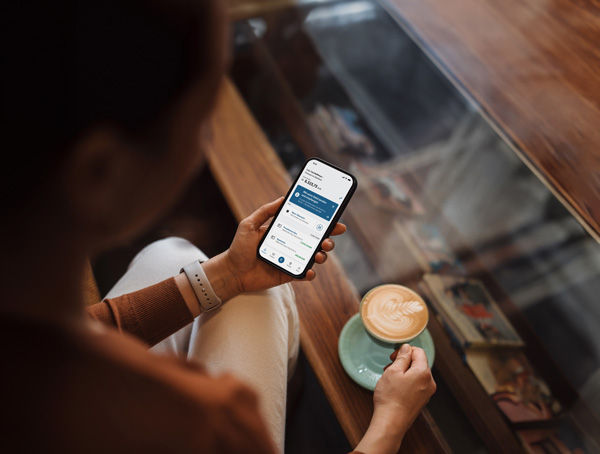 Frau mit einem Kaffe in der rechten und dem Handy mit geöffneter BBBank-BankingApp in der linken Hand