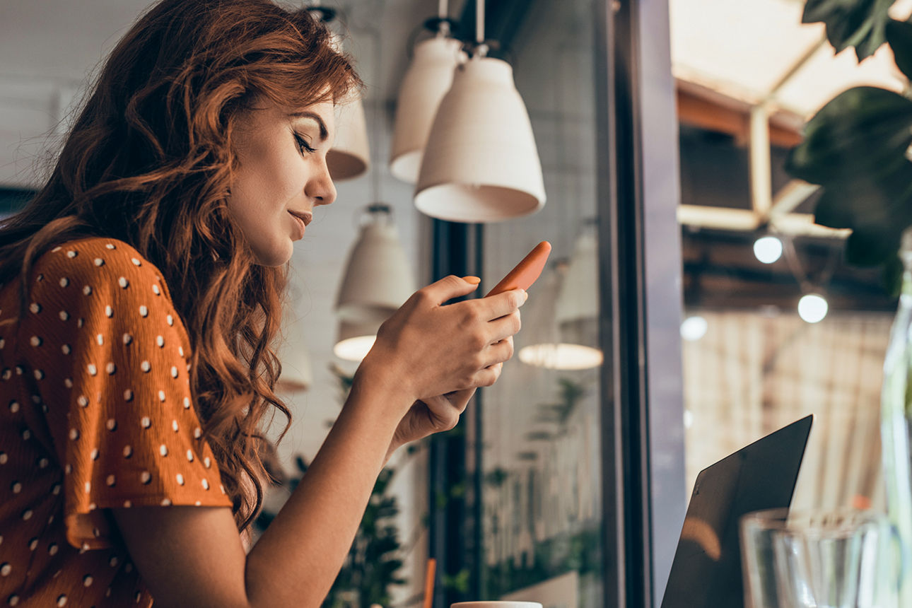 Frau mit rötlichen Haaren sitzt an einem Tisch im vor einem Laptop und schaut auf ihr Smartphone