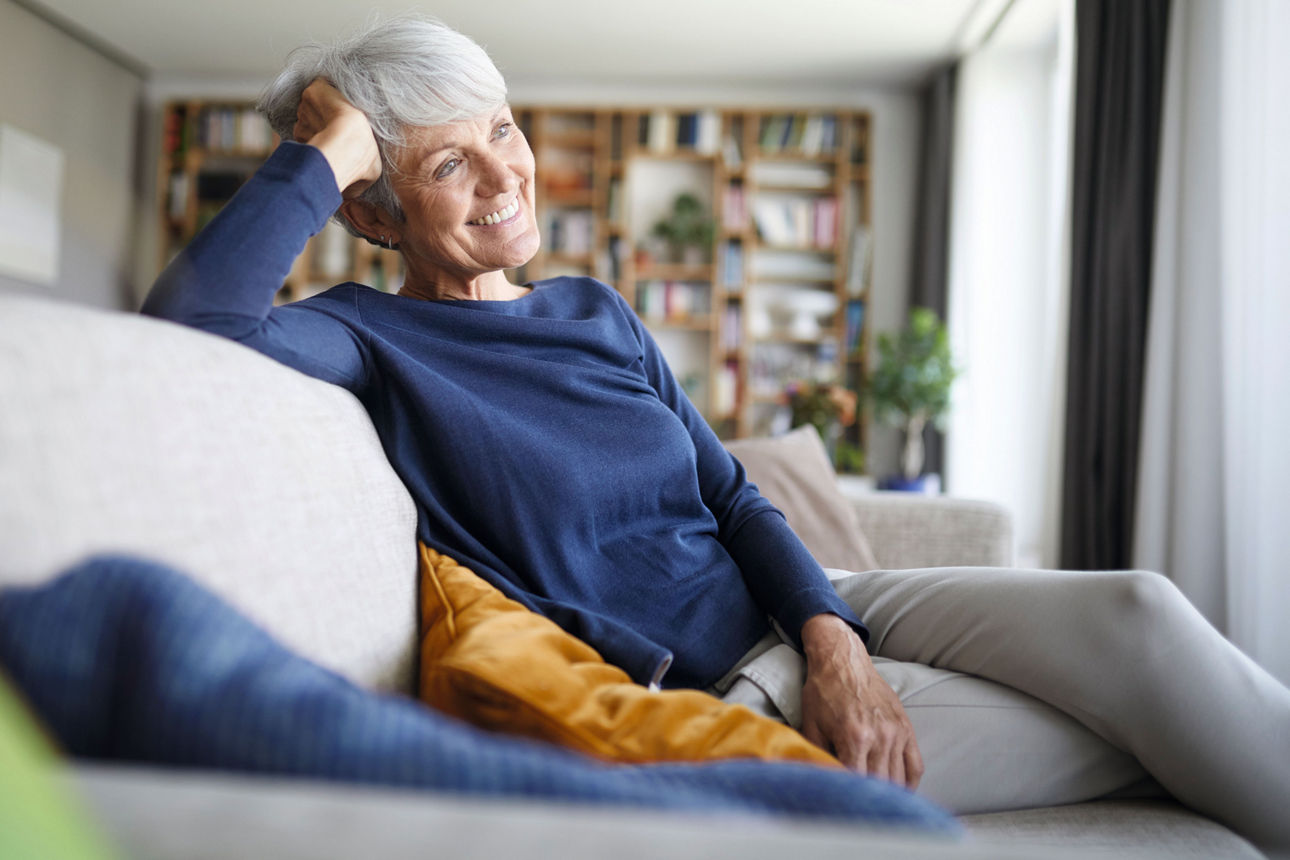 Person mit grauem Haar sitzt auf einem Sofa in einem hellen Wohnzimmer.