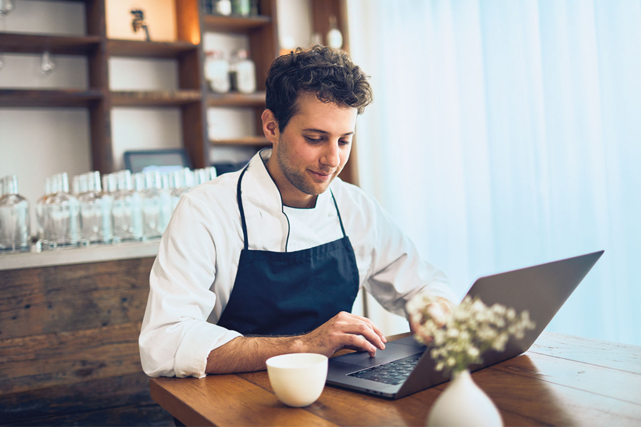 Koch in Schürze sitzt in einem Restaurant vor einem Laptop