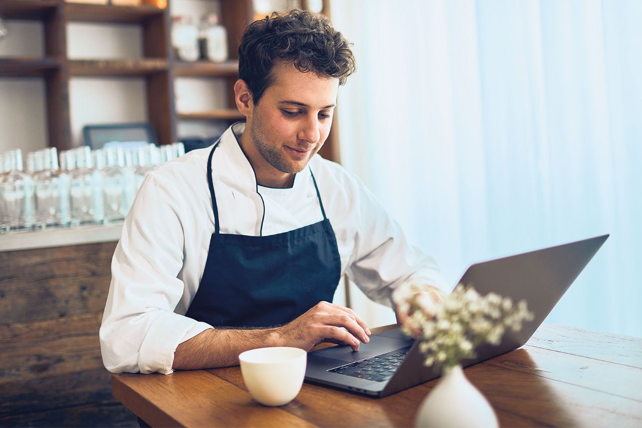 Koch in Schürze sitzt in einem Restaurant vor einem Laptop