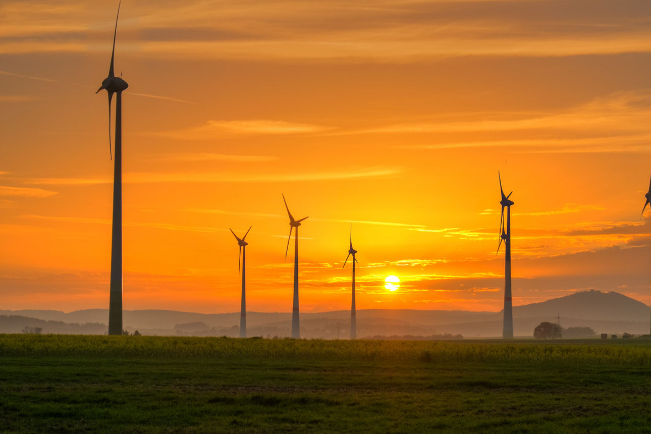Windräder stehen auf einem Feld, im Hintergrund sieht man Berge und den Sonnenuntergang.